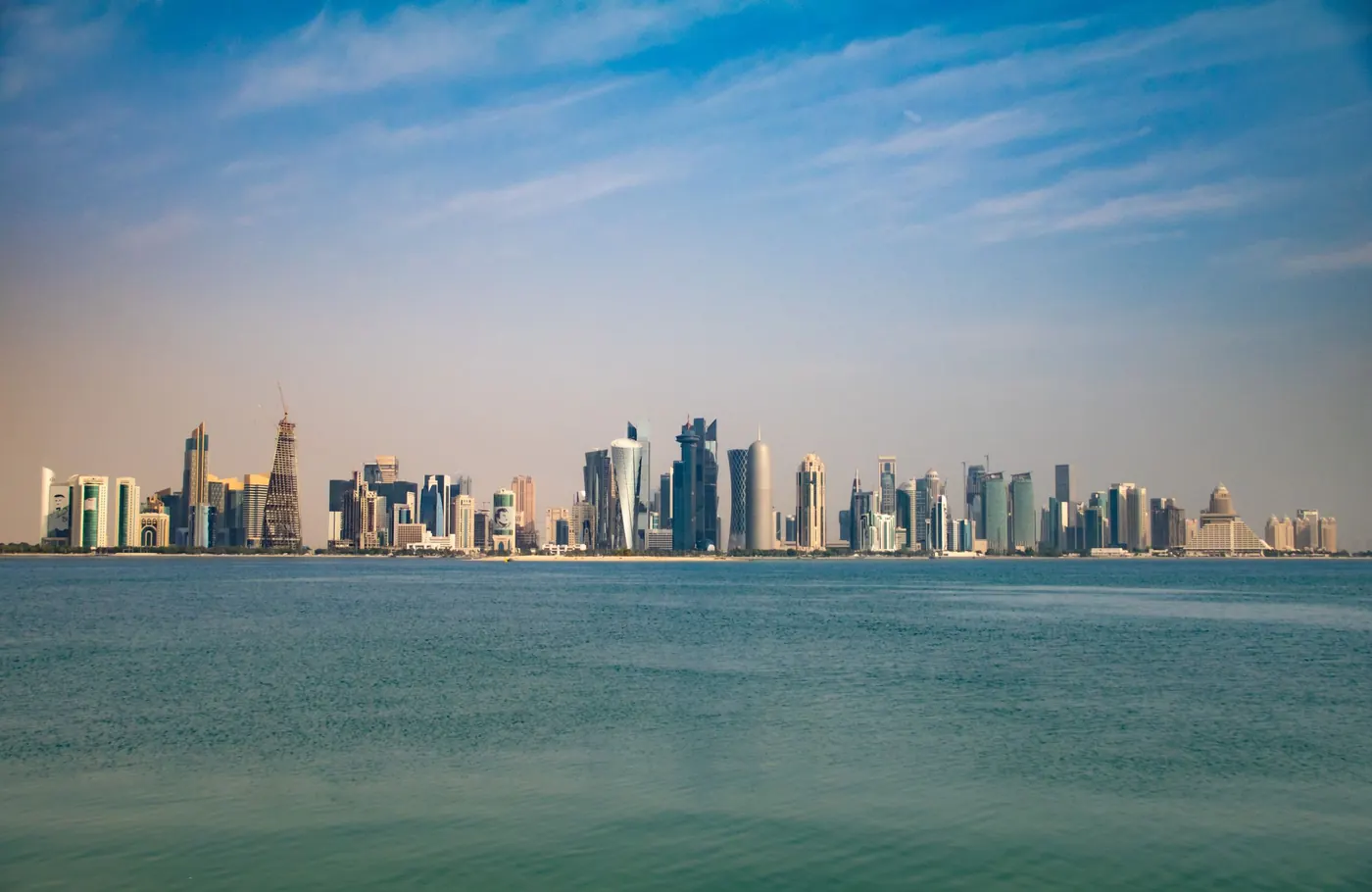 The Doha skyline across turquoise water on a clear day, seen from the Corniche
