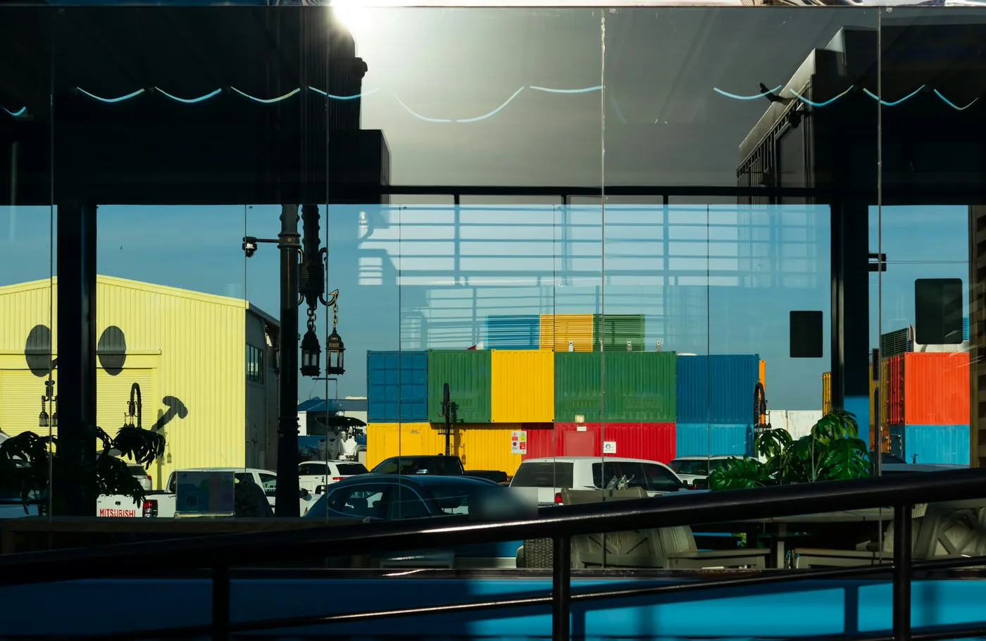 Stacked shipping containers in blue, green, yellow and red, seen through a glass window with parked cars in front