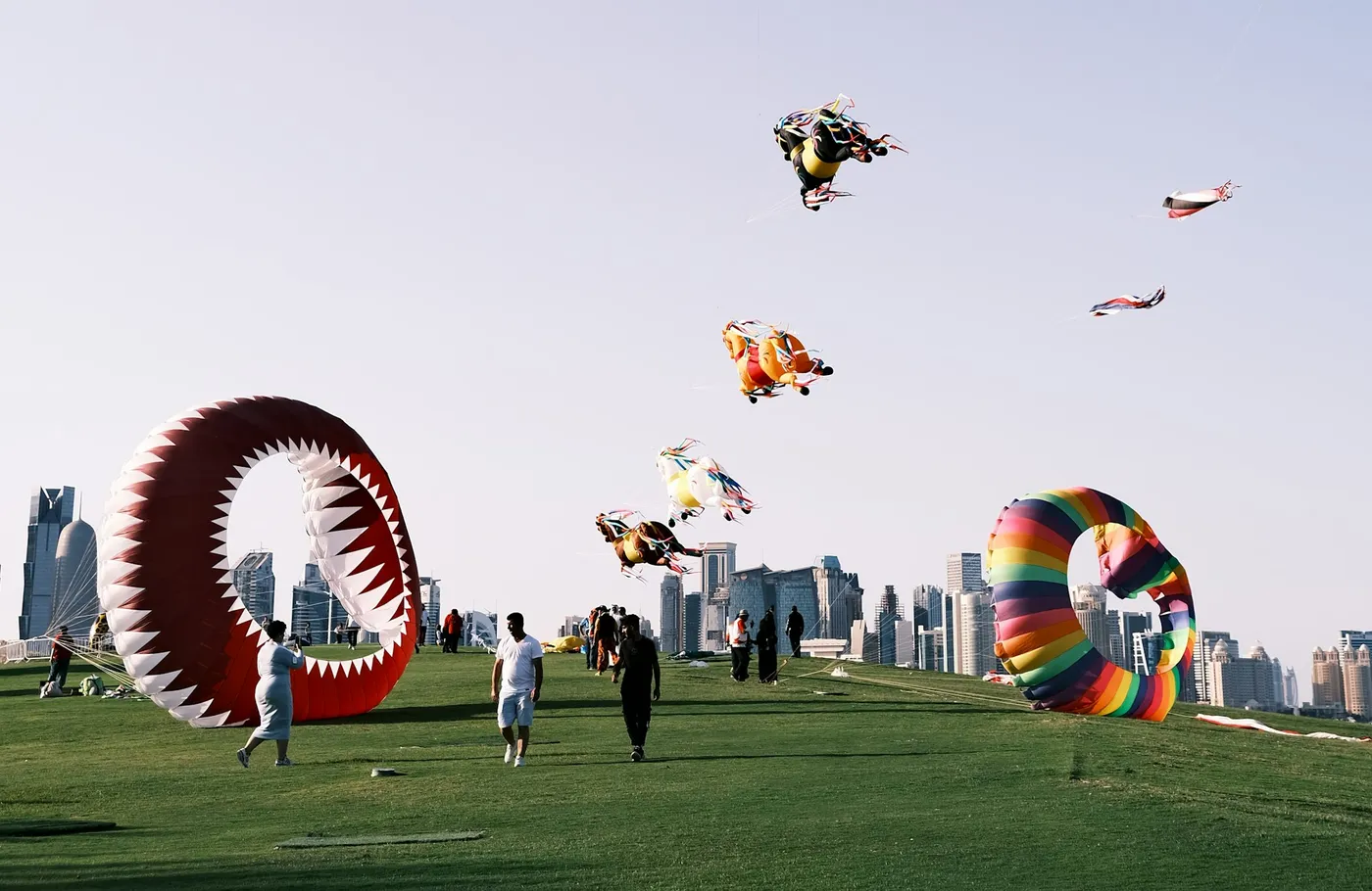People flying large kites on the lawn of MIA Park, with the West Bay skyline behind