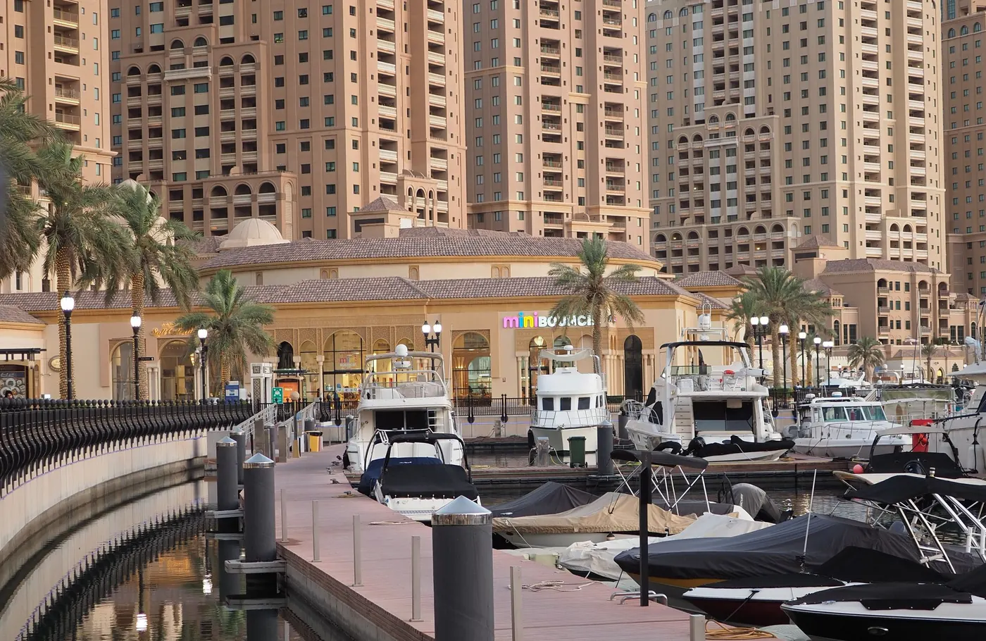 Boats moored along a marina promenade with palm trees and apartment towers behind