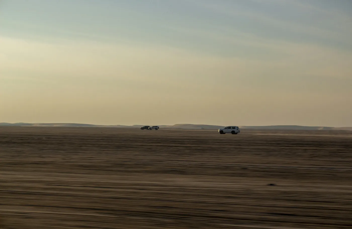 Three four wheel drives crossing flat desert towards low sand dunes, in evening light