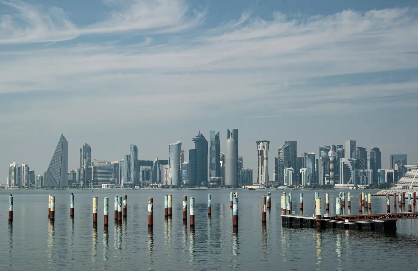 Doha’s West Bay skyline across calm water, with wooden posts in the foreground